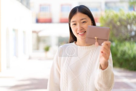 Téléchargez les photos : Jolie femme chinoise tenant un portefeuille à l'extérieur avec une expression heureuse - en image libre de droit