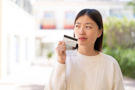 Foto de Mujer china bonita al aire libre sosteniendo una tarjeta de crédito y pensando - Imagen libre de derechos