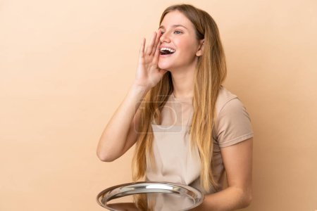 Foto de Joven camarera rubia con bandeja aislada sobre fondo beige gritando con la boca abierta hacia un lado - Imagen libre de derechos