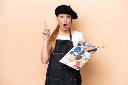 Téléchargez les photos : Jeune artiste femme caucasienne tenant une palette isolée sur fond beige dans le but de réaliser la solution tout en levant le doigt - en image libre de droit