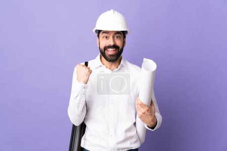 Photo for Young architect man with helmet and holding blueprints over isolated purple background celebrating a victory in winner position - Royalty Free Image