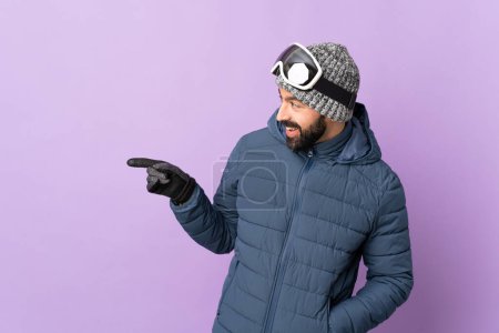 Téléchargez les photos : Skier homme avec des lunettes de snowboard sur fond violet isolé pointant du doigt sur le côté et présentant un produit - en image libre de droit