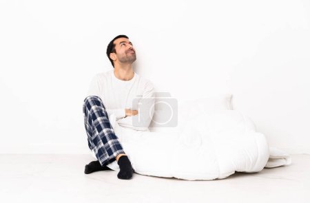 Téléchargez les photos : Homme caucasien en pyjama assis sur le sol à l'intérieur les yeux levés tout en souriant - en image libre de droit