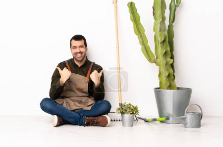 Photo for Gardener man sitting on the floor at indoors with thumbs up gesture and smiling - Royalty Free Image