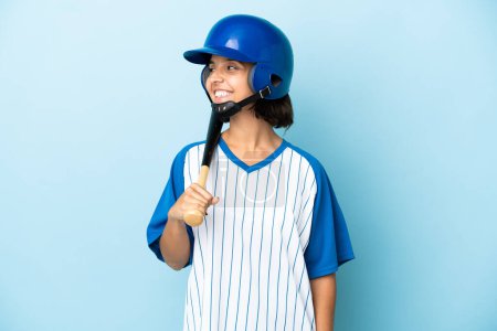 Téléchargez les photos : Baseball mixte femme joueuse avec casque et chauve-souris isolé sur fond bleu regardant côté - en image libre de droit