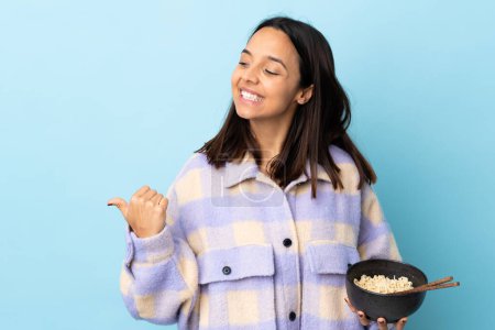 Photo for Young brunette mixed race woman over isolated blue background pointing to the side to present a product while holding a bowl of noodles with chopsticks. - Royalty Free Image