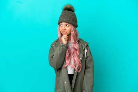 Junge Frau mit pinkfarbenen Haaren, die einen regenfesten Mantel auf blauem Hintergrund trägt und beim Nachdenken eine Idee hat