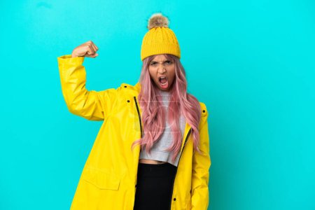 Young woman with pink hair wearing a rainproof coat isolated on blue background doing strong gesture