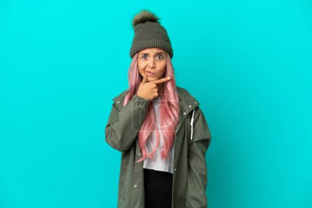 Junge Frau mit pinkfarbenen Haaren, die einen regenfesten Mantel auf blauem Hintergrund trägt, hat Zweifel