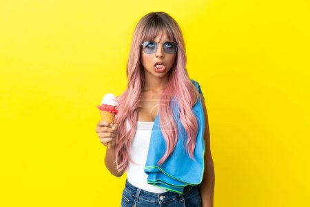 Foto de Mujer joven de raza mixta con cabello rosa sosteniendo helado aislado sobre fondo amarillo mirando hacia arriba y con expresión sorprendida - Imagen libre de derechos