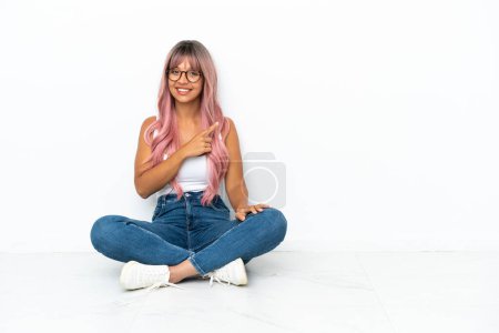Foto de Mujer joven de raza mixta con pelo rosa sentada en el suelo aislada sobre fondo blanco apuntando hacia un lado para presentar un producto - Imagen libre de derechos