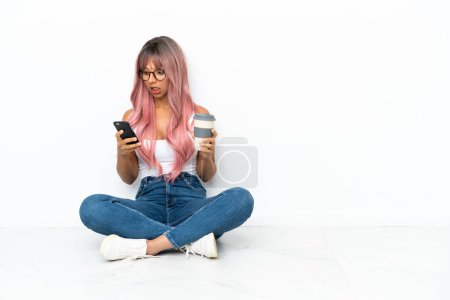 Téléchargez les photos : Jeune femme métisse aux cheveux roses assise sur le sol isolé sur fond blanc tenant du café à emporter et un mobile - en image libre de droit