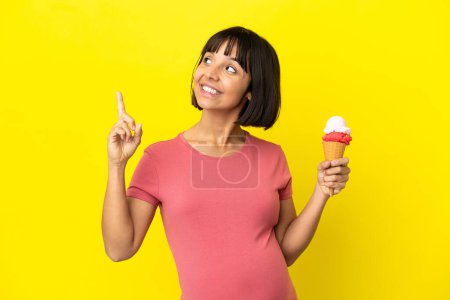 Foto de Mujer embarazada sosteniendo un helado de corneta aislado sobre fondo amarillo señalando una gran idea - Imagen libre de derechos