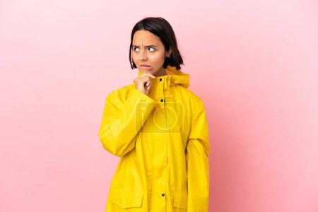 Junge lateinamerikanische Frau trägt einen regendichten Mantel über isoliertem Hintergrund, hat Zweifel und denkt nach