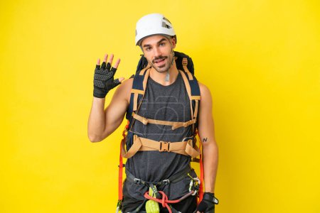 Foto de Joven escalador caucásico aislado sobre fondo amarillo saludando con la mano con expresión feliz - Imagen libre de derechos