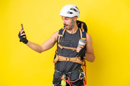 Young caucasian rock climber man isolated on yellow background holding coffee to take away and a mobile