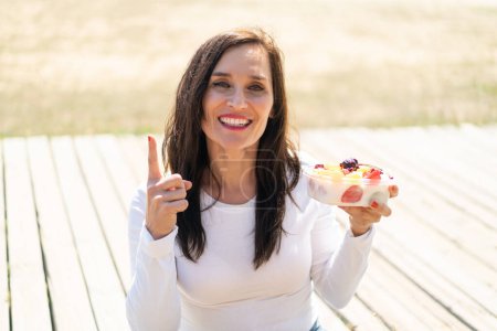 Téléchargez les photos : Femme d'âge moyen tenant un bol de fruits à l'extérieur pointant vers le haut d'une grande idée - en image libre de droit