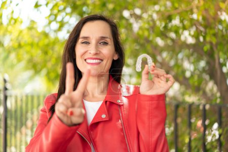 Téléchargez les photos : Femme d'âge moyen tenant des bretelles invisibles à l'extérieur souriant et montrant signe de victoire - en image libre de droit