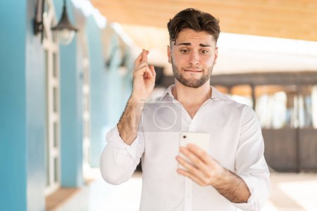 Photo for Young handsome man using mobile phone with fingers crossing - Royalty Free Image