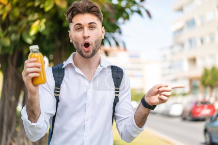 Téléchargez les photos : Jeune homme beau tenant un jus d'orange à l'extérieur surpris et pointant côté - en image libre de droit