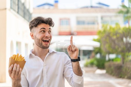 Téléchargez les photos : Jeune homme beau tenant des chips frites à l'extérieur dans le but de réaliser la solution tout en levant le doigt - en image libre de droit