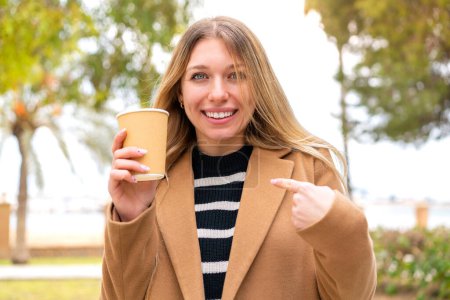 Foto de Joven mujer rubia bonita sosteniendo un café para llevar al aire libre con expresión facial sorpresa - Imagen libre de derechos