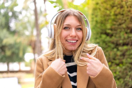 Téléchargez les photos : Jeune jolie femme blonde écoutant de la musique avec des écouteurs à l'extérieur avec une expression faciale surprise - en image libre de droit