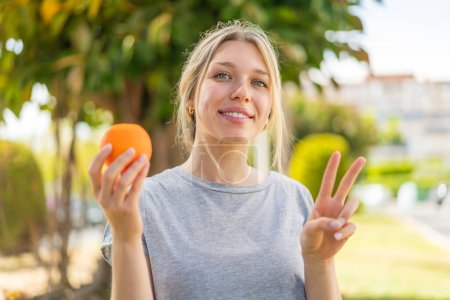 Foto de Mujer rubia joven sosteniendo una naranja al aire libre sonriendo y mostrando signo de victoria - Imagen libre de derechos