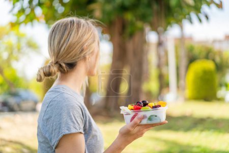 Téléchargez les photos : Jeune femme blonde tenant un bol de fruits à l'extérieur en position arrière - en image libre de droit
