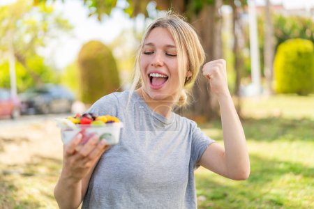 Téléchargez les photos : Jeune femme blonde tenant un bol de fruits à l'extérieur célébrant une victoire - en image libre de droit