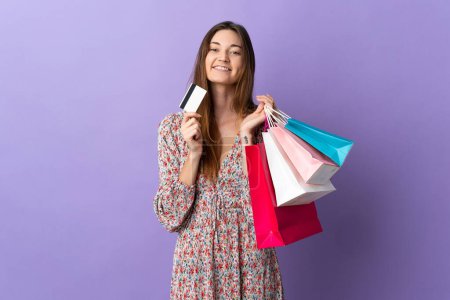 Foto de Mujer joven de Irlanda aislada sobre fondo púrpura sosteniendo bolsas de compras y una tarjeta de crédito y pensando - Imagen libre de derechos