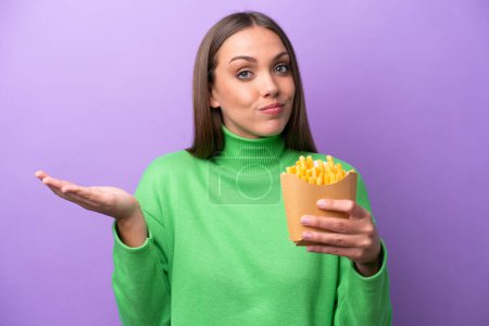 Photo for Young caucasian woman holding fried chips on purple background having doubts while raising hands - Royalty Free Image