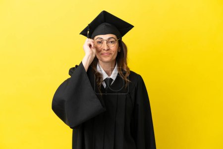 Foto de Graduado universitario de mediana edad aislado sobre fondo amarillo con gafas y sorprendido - Imagen libre de derechos