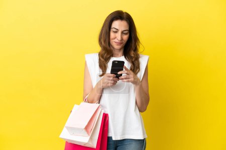 Photo for Middle aged caucasian woman isolated on yellow background holding shopping bags and writing a message with her cell phone to a friend - Royalty Free Image