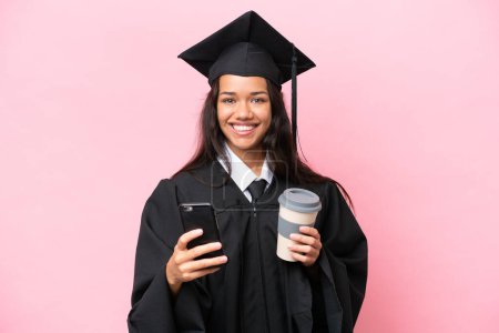 Téléchargez les photos : Jeune universitaire colombienne diplômée isolée sur fond rose tenant du café à emporter et un mobile - en image libre de droit