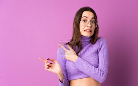Photo for Young woman over isolated purple background frightened and pointing to the side - Royalty Free Image