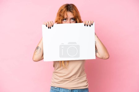Téléchargez les photos : Jeune femme caucasienne isolée sur fond rose tenant une pancarte vide et se cachant derrière elle - en image libre de droit