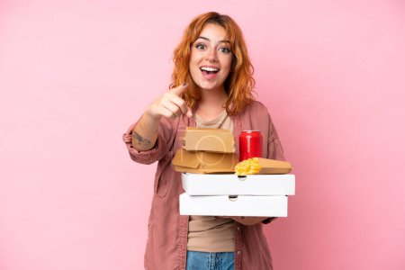 Photo for Young caucasian woman holding fast food isolated on pink background surprised and pointing front - Royalty Free Image