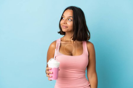 Photo for Young woman with strawberry milkshake isolated on blue background having doubts while looking side - Royalty Free Image