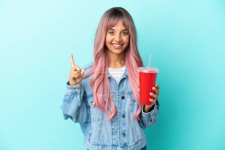 Foto de Joven mujer de raza mixta bebiendo una bebida fresca aislada sobre fondo azul señalando una gran idea - Imagen libre de derechos