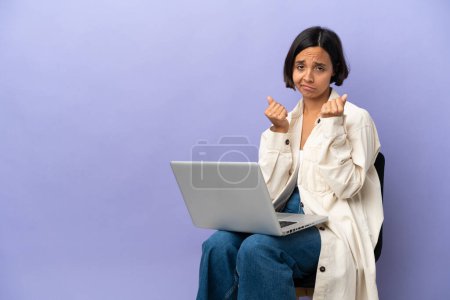 Junge gemischte Rasse Frau sitzt auf einem Stuhl mit Laptop isoliert auf lila Hintergrund macht Geld Geste, ist aber ruiniert