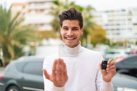 Téléchargez les photos : Jeune homme caucasien tenant des clés de voiture à l'extérieur invitant à venir avec la main. Heureux que tu sois venu - en image libre de droit