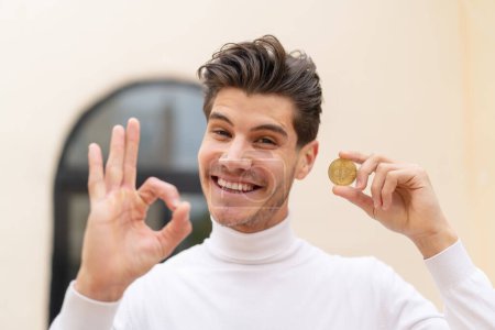 Photo for Young caucasian man holding a Bitcoin at outdoors showing ok sign with fingers - Royalty Free Image