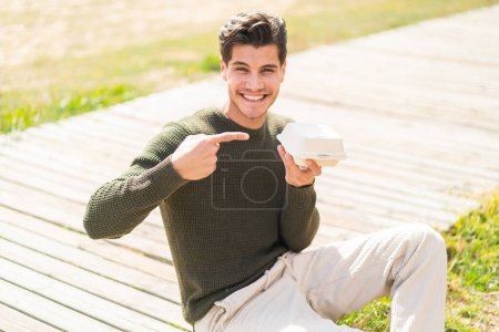 Photo for Young caucasian man at outdoors taking a box of takeaway food and pointing it - Royalty Free Image
