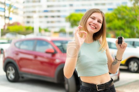 Téléchargez les photos : Jeune femme blonde tenant des clés de voiture à l'extérieur montrant ok signe avec les doigts - en image libre de droit