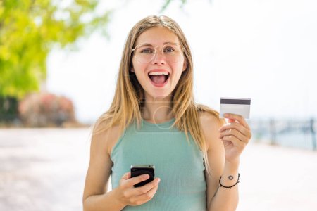 Foto de Joven mujer rubia al aire libre comprando con el móvil y sosteniendo una tarjeta de crédito con expresión sorprendida - Imagen libre de derechos
