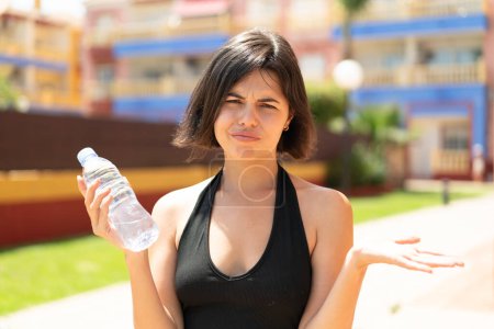 Foto de Joven bonita mujer búlgara con una botella de agua al aire libre haciendo gestos de dudas mientras levanta los hombros - Imagen libre de derechos