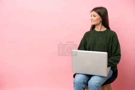 Photo for Young caucasian woman sitting on a chair with her laptop isolated on pink background looking side - Royalty Free Image