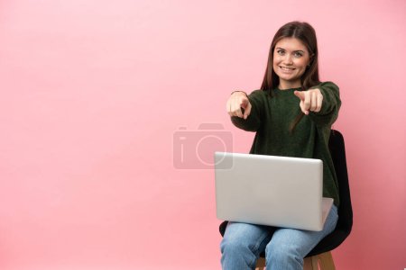 Téléchargez les photos : Jeune femme caucasienne assise sur une chaise avec son ordinateur portable isolé sur fond rose pointe doigt à vous tout en souriant - en image libre de droit
