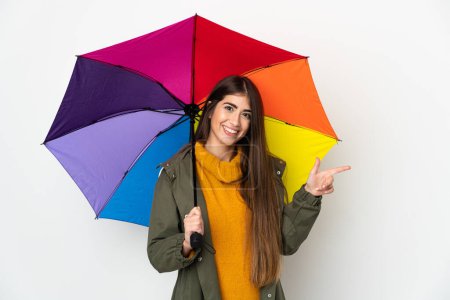 Téléchargez les photos : Jeune femme tenant un parapluie isolé sur fond blanc pointant du doigt sur le côté - en image libre de droit